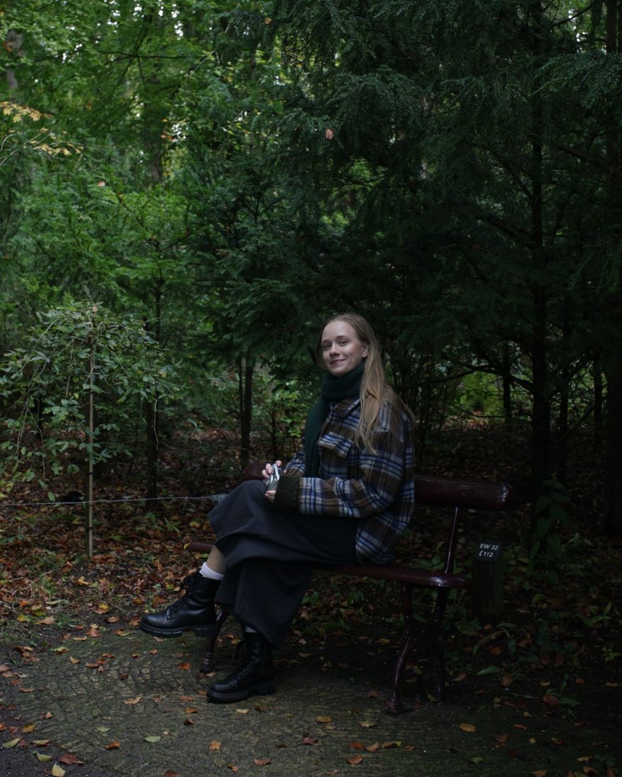 Zsófi sitting on a bench in an autumn forest, holding a film camera